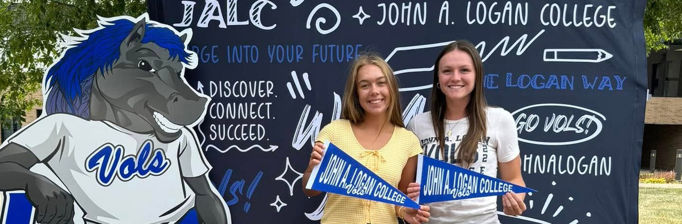 Two students holding John A. Logan College Vols pennants next to a cutout of JALC's mascot Charger.